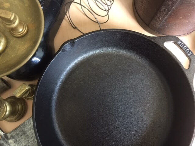 Why use a cast iron skillet? In the picture a Lodge skillet is on a wooden table. Also in the table are old fashioned cooking utensils.