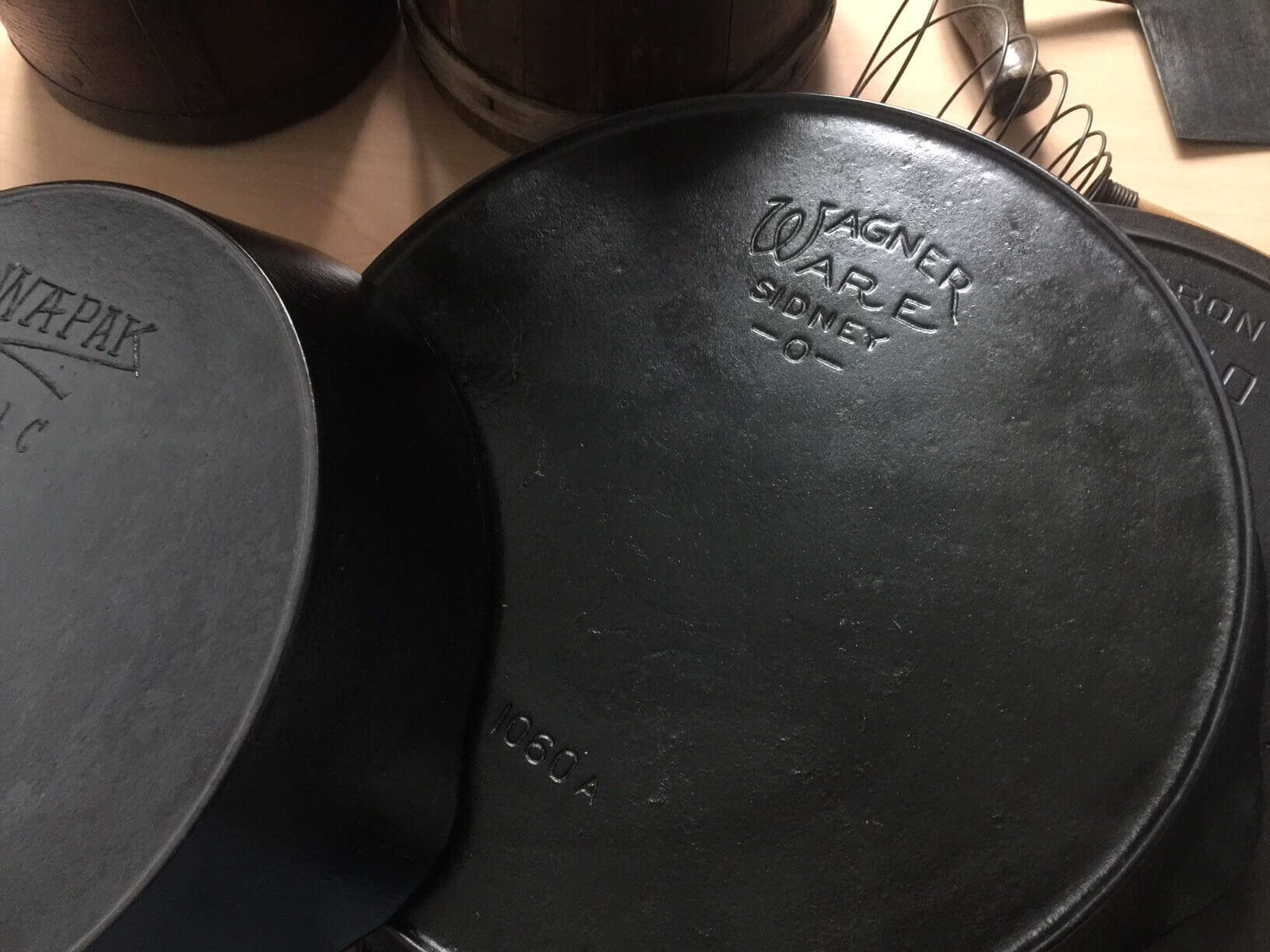 Three cast iron skillets on a table. on the left is a Wapak in the middle Wagner and the is a Griswold.